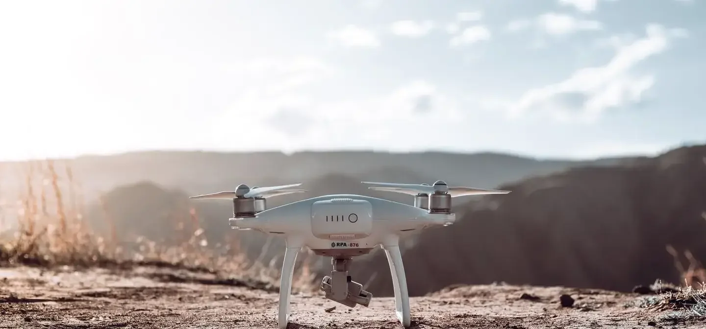Offene Kategorie Drohne Regeln - Drohnen Führerschein Deutschland Front view of a white quadcopter drone on a dry, rocky ground with sunlit hills in the distance at sunset/late afternoon.